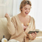 Woman using Medela Swing Maxi hands-free double breast pump while sitting on a couch and smiling at her phone. The pump fits securely inside her bra, freeing her hands for daily tasks.