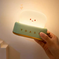 Hand holding green toaster night light by the brass lever showing portable size and warm glowing toast lamp face
