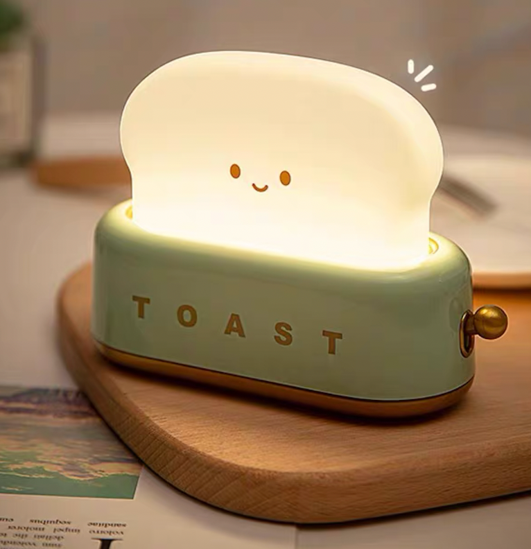 Green toaster night light on wooden chopping board with cutlery, lit toast slice with smiley face emitting warm light